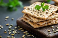 Crispy multigrain crackers with pumpkin seeds stacked on wooden board, garnished with fresh herbs. Close up view of healthy snack with selective focus.