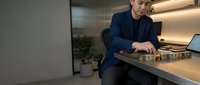 Young Asian businessman in dark suit working late at modern office desk with laptop and documents in contemporary workspace with warm lighting.
