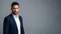 Adult male with dark hair and facial stubble, Mediterranean ethnicity, wearing navy blazer over white shirt. Corporate portrait against neutral gray background for business and professional use.