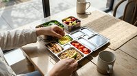 Person in casual beige sweater organizing meal prep containers with fresh vegetables, grains and proteins on wooden table for weekly food planning and healthy eating routine.