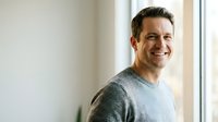 Happy middle-aged Caucasian man in gray sweater smiling warmly by window in modern home interior for lifestyle and wellness content.