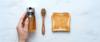 Hand holding honey jar with wooden dipper and golden toast on marble surface, breakfast ingredients for cooking and food preparation.