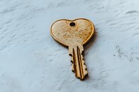 Single brass house key with heart-shaped bow lying on textured white marble surface, showing wear and patina from regular use. Macro photography captures detail.