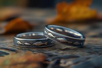 Pair of silver wedding bands with engraved wave pattern on rustic wooden surface with autumn leaves, capturing intimate matrimonial symbolism in warm natural lighting.