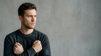 Young adult male with dark hair and light skin tone wearing dark gray sweater, looking away with confident expression against neutral gray background for portrait use.