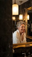 Happy mature Caucasian woman with gray hair in white bathrobe looking at mirror reflection in luxury bathroom with pendant lights and marble walls.
