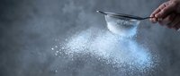 Hand sifting white flour through metal strainer creating powder cloud against dark background for baking and cooking preparation.