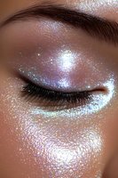 Closed eye with shimmering holographic glitter makeup in iridescent pearl tones, featuring dramatic black lashes and defined brow on medium skin tone.