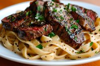 Juicy grilled steak slices served on creamy fettuccine pasta garnished with fresh herbs and black pepper, creating a luxurious main course presentation.