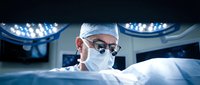 Male surgeon in surgical mask and cap performing operation under bright medical lights in modern operating room for healthcare and medical procedure concepts.