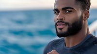 Young African American man with beard smiling confidently against blurred ocean background, wearing navy blue casual shirt. Natural outdoor portrait capturing genuine expression.