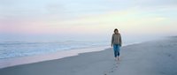 Solitary person walking along empty beach at sunset with soft pink and purple sky colors reflecting on wet sand and gentle ocean waves.