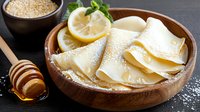 Thin crepes with honey drizzle and lemon slices served in wooden bowl, garnished with fresh mint leaves and powdered sugar, sesame seeds aside.
