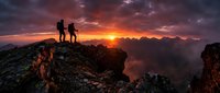 Silhouettes of hikers standing on rocky mountain peak at dramatic sunset with golden sky and distant mountain range, adventure and outdoor exploration concept.