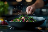 Fresh mixed greens salad with herbs being seasoned by chef hand sprinkling salt in dark bowl on kitchen counter for healthy dining and culinary preparation.