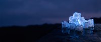 Pile of translucent ice cubes stacked on wet surface against dark blue night sky background, suitable for beverage, cooling and refreshment concepts.