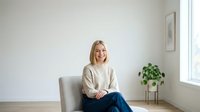 Young woman with blonde hair, Caucasian ethnicity, wearing casual beige cardigan and jeans, sitting relaxed on gray armchair in bright minimalist interior with potted plant and window light.