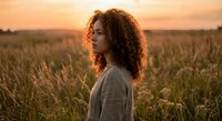 Young mixed-race woman with curly hair standing in golden wheat field during sunset, peaceful countryside moment for wellness and lifestyle content.