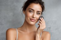 Young Caucasian woman with natural makeup applying facial skincare product against gray concrete background, showcasing beauty routine.