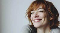 Candid portrait of young redhead woman with genuine smile and natural freckles wearing casual grey sweater against light background. Positive emotional expression.