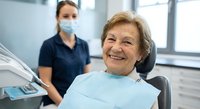 Happy elderly Caucasian woman patient smiling in dental chair with masked female dentist in background at modern clinic for oral healthcare checkup.