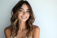 Young woman with radiant smile and wavy brown highlighted hair against light background, wearing natural makeup and showing genuine happiness in casual portrait.