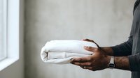 Male hands with dark skin tone holding folded white linens or towels indoors near a window, suitable for hospitality, laundry, and household management concepts.