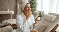 Young woman in white bathrobe and towel applying skincare cream in bright bedroom. Morning beauty routine, self-care wellness lifestyle concept.
