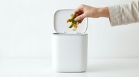 Hand dropping vegetable peels into a white compost bin on a clean surface, promoting sustainable kitchen waste management and eco-friendly living.