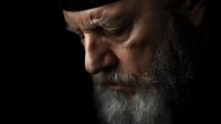 Dramatic side profile portrait of senior bearded man with intense gaze and weathered features against dark background, captured in moody low-key lighting.