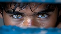 Intense close-up of piercing amber eyes with dark eyebrows and tanned skin, partially obscured by blue fabric creating dramatic lighting and mysterious atmosphere.