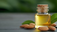 Golden almond oil in glass bottle with cork stopper surrounded by fresh almonds and green leaves on wooden surface for natural skincare and aromatherapy.