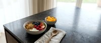 Overhead view of a breakfast bowl with granola, berries, and yogurt on a dark countertop, accompanied by fresh fruit and utensils for morning meal preparation and healthy eating content.