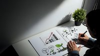 Business professional drawing growth strategy diagrams with charts, lightbulbs and money symbols on white paper at modern office desk with plant.