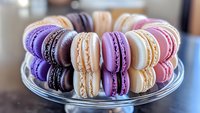 Colorful French macarons arranged on glass plate with purple, white, pink and orange varieties showing delicate meringue texture and cream filling. Sweet dessert display.
