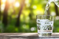 Fresh water pouring into clear glass on wooden surface against blurred green nature background with sunlight, showcasing purity and refreshment.