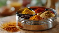 Vibrant aromatic spices in traditional metal Indian masala box with turmeric, paprika, cumin and curry powder scattered on wooden surface, soft natural lighting.