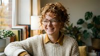 Young curly-haired woman with glasses smiling warmly in cozy home office with plants, natural lighting for lifestyle and remote work concepts.