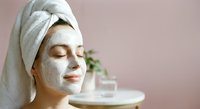 Young woman with white clay facial mask and towel on head relaxing during spa skincare routine at home with closed eyes and serene expression.