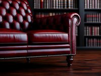 Luxurious burgundy leather Chesterfield sofa with classic tufted upholstery and nailhead trim against dark library bookshelf background on hardwood floor.