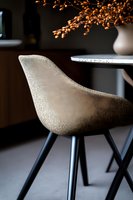 Modern beige upholstered dining chair with black metal legs against dark background, elegant round table and decorative dried flowers in white vase create cozy atmosphere.