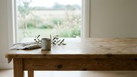 Wooden table with small vase of olive branches near bright window overlooking countryside landscape, minimalist interior styling for home decor and design projects.