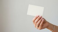 Woman's hand holding blank white card against neutral background, displaying manicured nails and jewelry for business, marketing, or promotional use.
