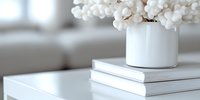White cotton stems arranged in ceramic vase on stack of books against blurred bright interior background, creating minimal modern home decor composition.
