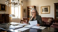 Mature Caucasian businesswoman with glasses reviewing documents in elegant office with chandelier and classic furniture.