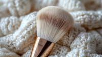 Luxurious makeup brush with rose gold handle resting on soft white knitted texture, showcasing professional beauty tool for cosmetic application and skincare routines.