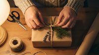 Young adult with medium skin tone wrapping kraft paper gift box with twine and fresh evergreen sprigs, surrounded by scissors, jute string and craft supplies on wooden table.