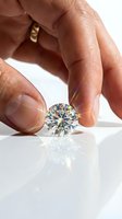 Hand holding a sparkling round brilliant cut diamond with rainbow light refractions on white reflective surface, luxury gemstone concept.