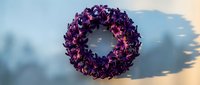 Purple dried flower wreath with natural petals arranged in circular pattern casting shadow on white surface for home decor and craft projects.