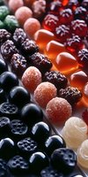 Colorful assortment of gummy candies and fruit jellies arranged in rows, including dark berries, sugar-coated pink spheres, and translucent orange pieces on white surface.